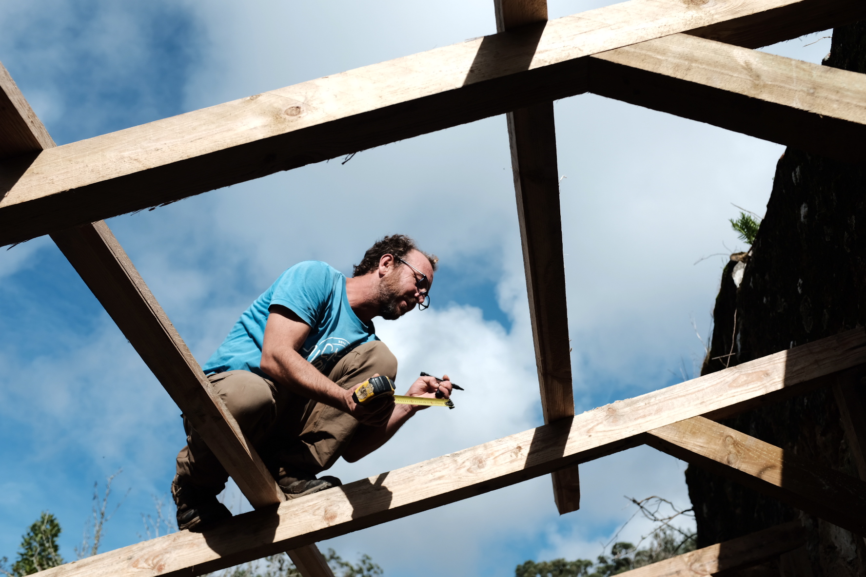 A man crouching on the roof framing with a tape measure against a blue sky
