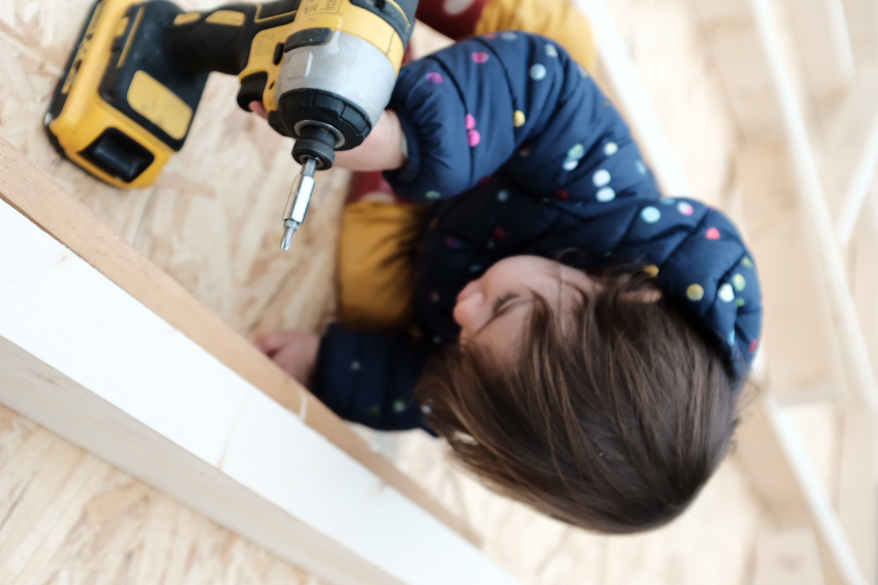A toddler in a spotted coat using a cordless drill on the floor deck