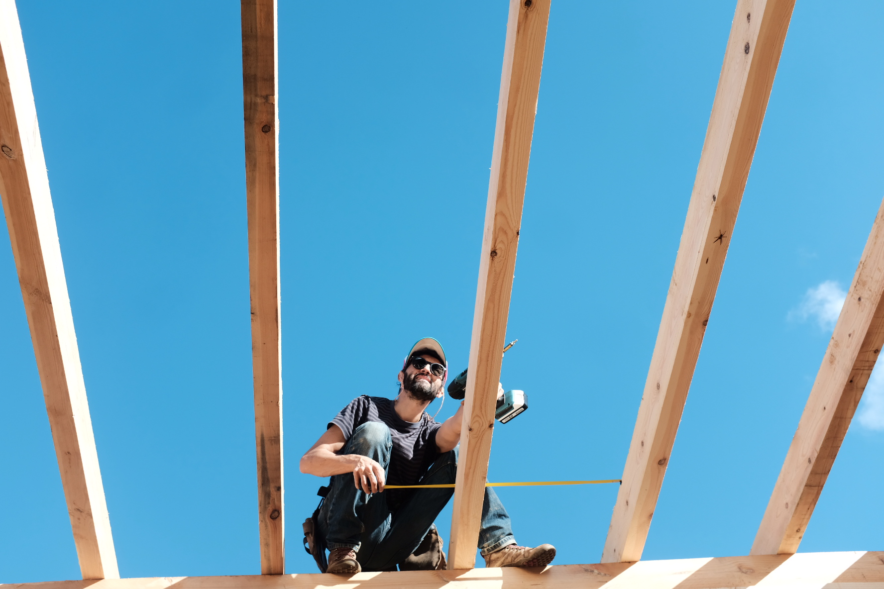A man crouching on the roof rafters with a tape measure against a blue sky