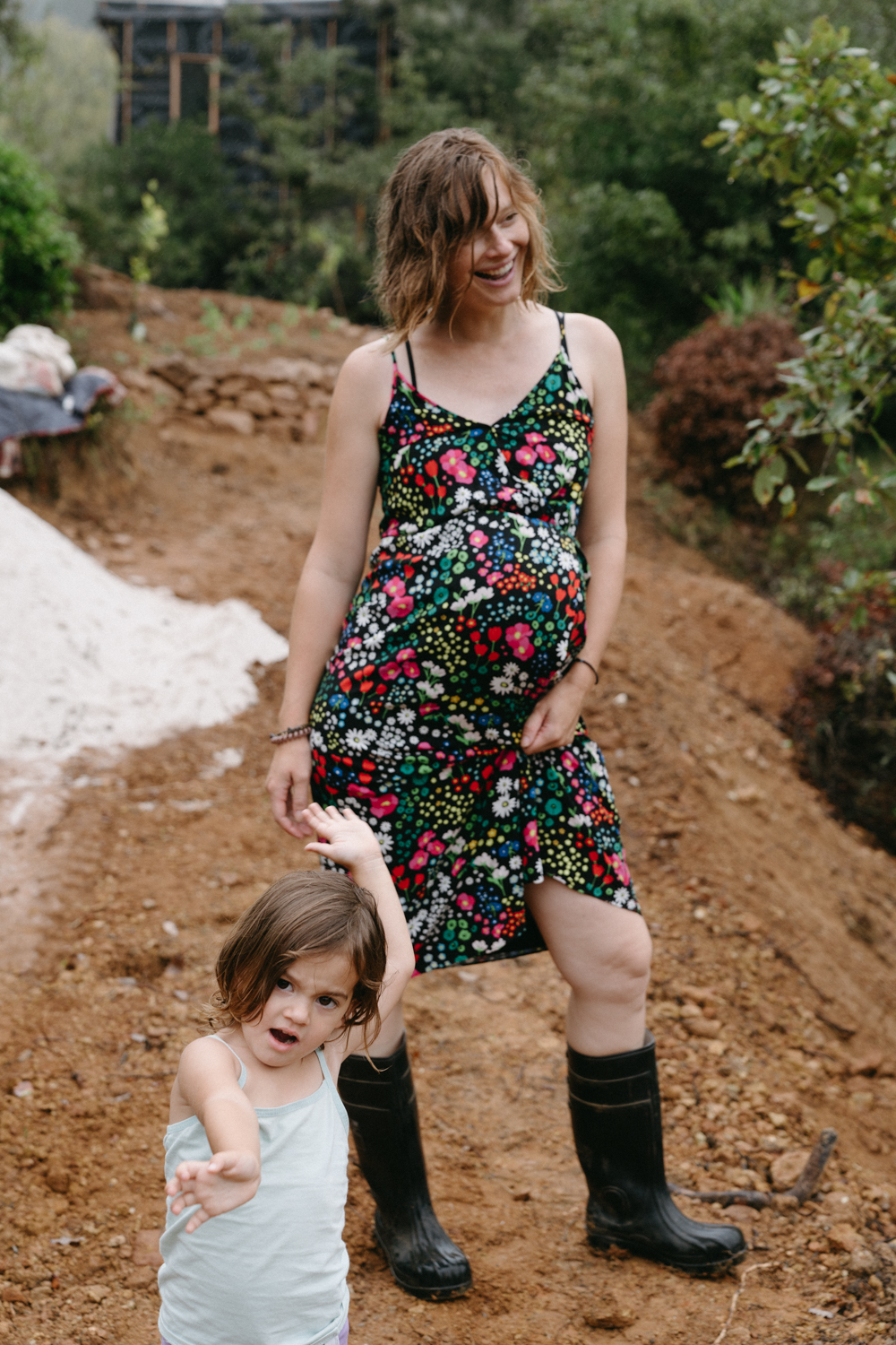 A pregnant woman in a floral dress and boots holding her daughter's hand on the bare earth site