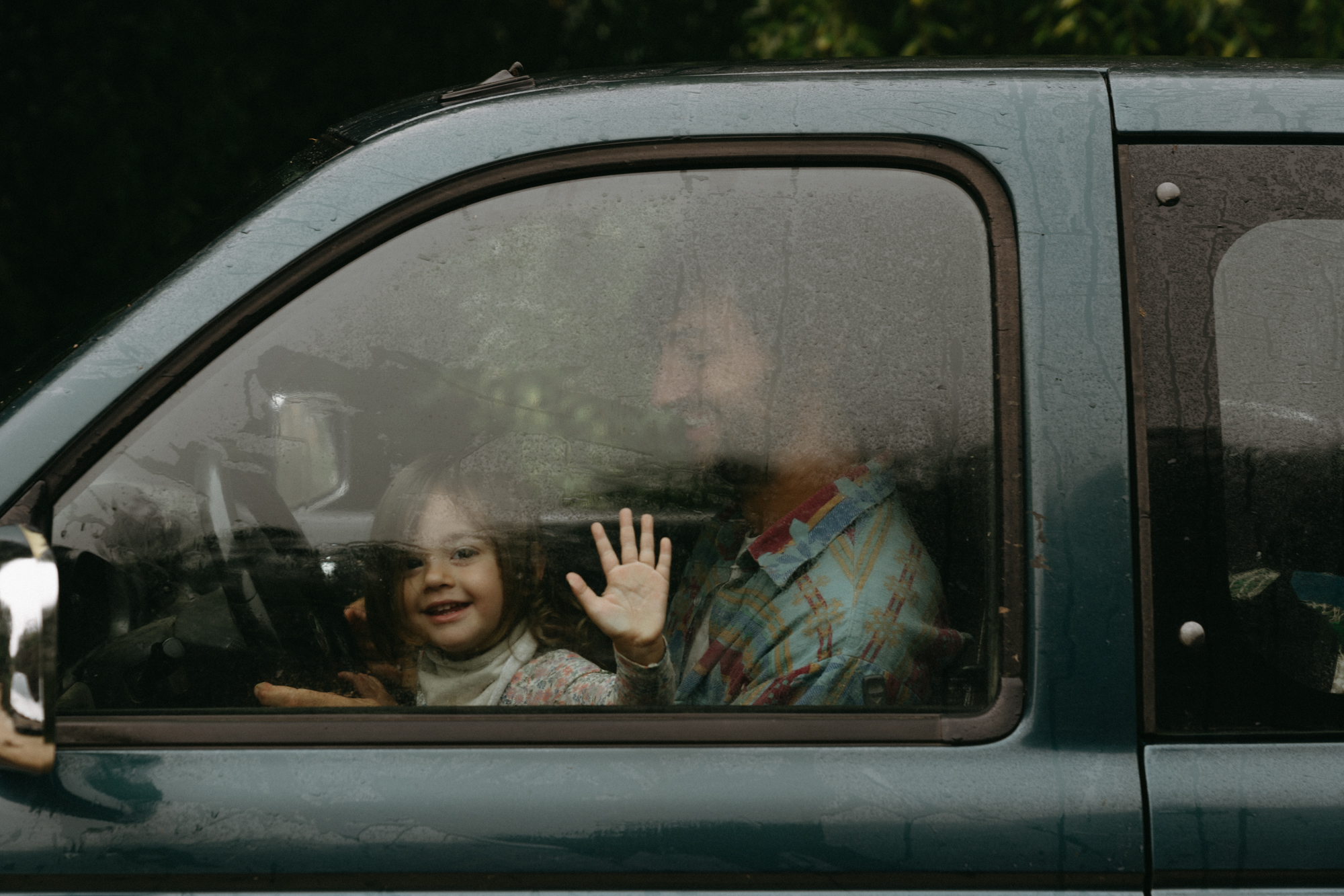 A man and a small girl waving from inside a rain-covered car window