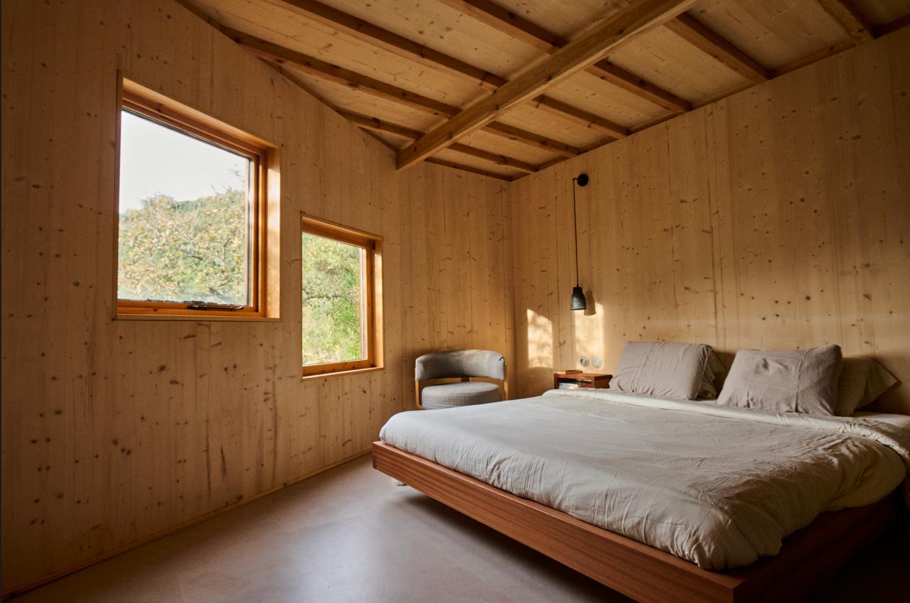 Timber bedroom with exposed rafters and two square windows onto the orchard