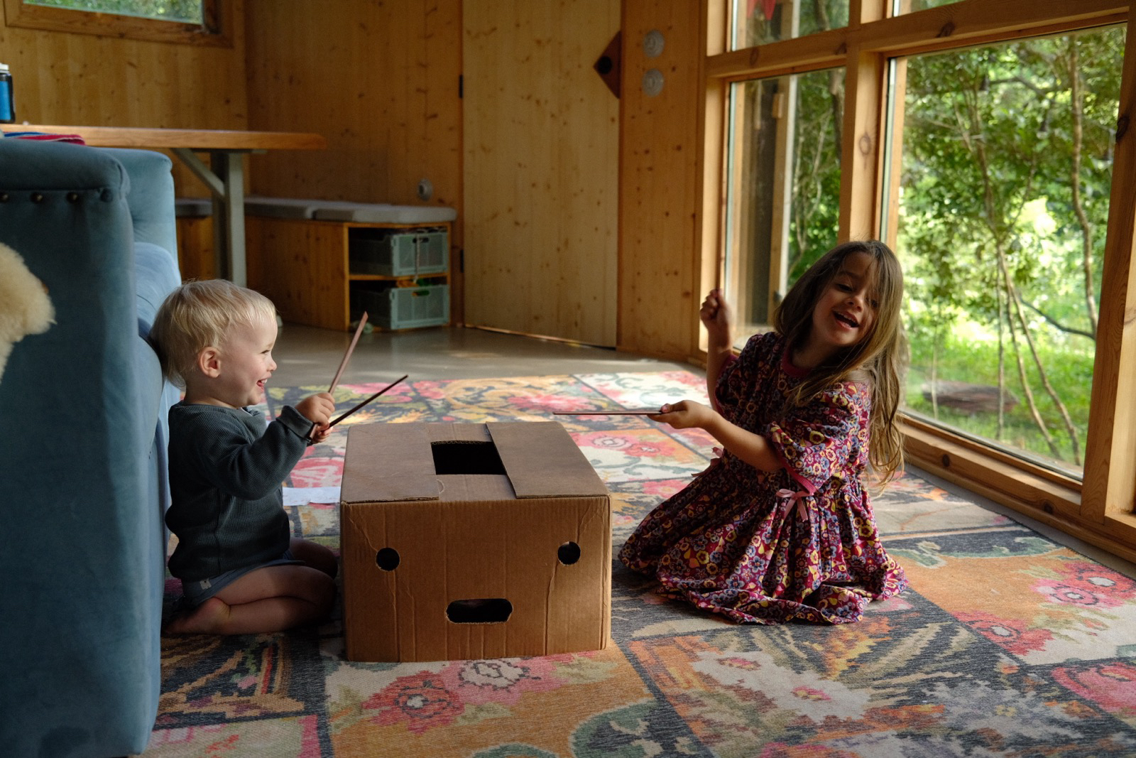 Two children drumming on a cardboard box on the rug in the timber living room