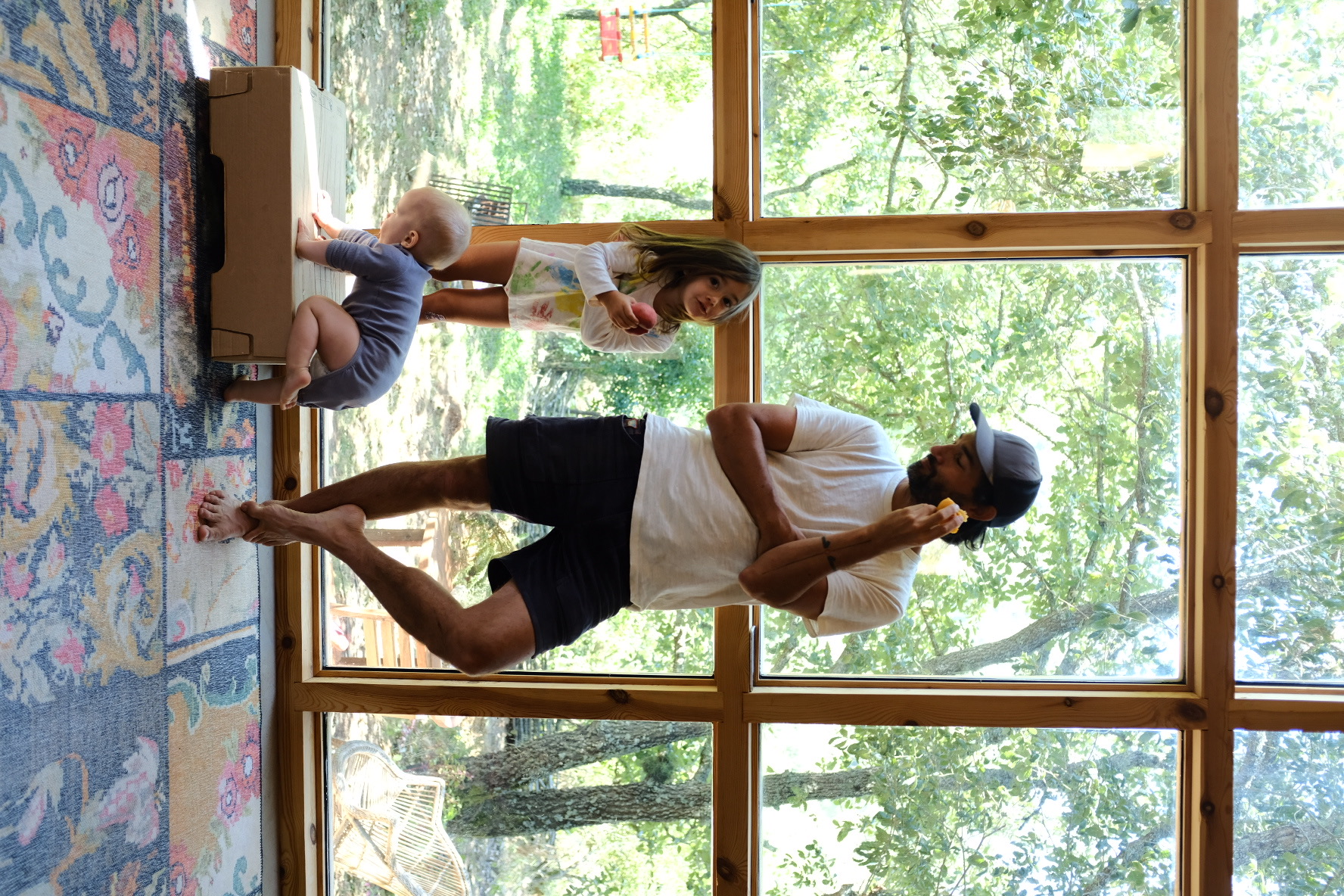 A man and his two children by the tall timber window wall, eating fruit